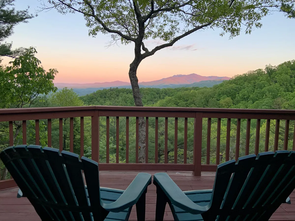 This image features a pair of Adirondack chairs on a wooden deck, perfectly positioned to take in sweeping mountain views. A tree in the foreground adds a touch of intimacy, while the expansive sky and distant ridgelines evoke the vastness of Pisgah National Forest.