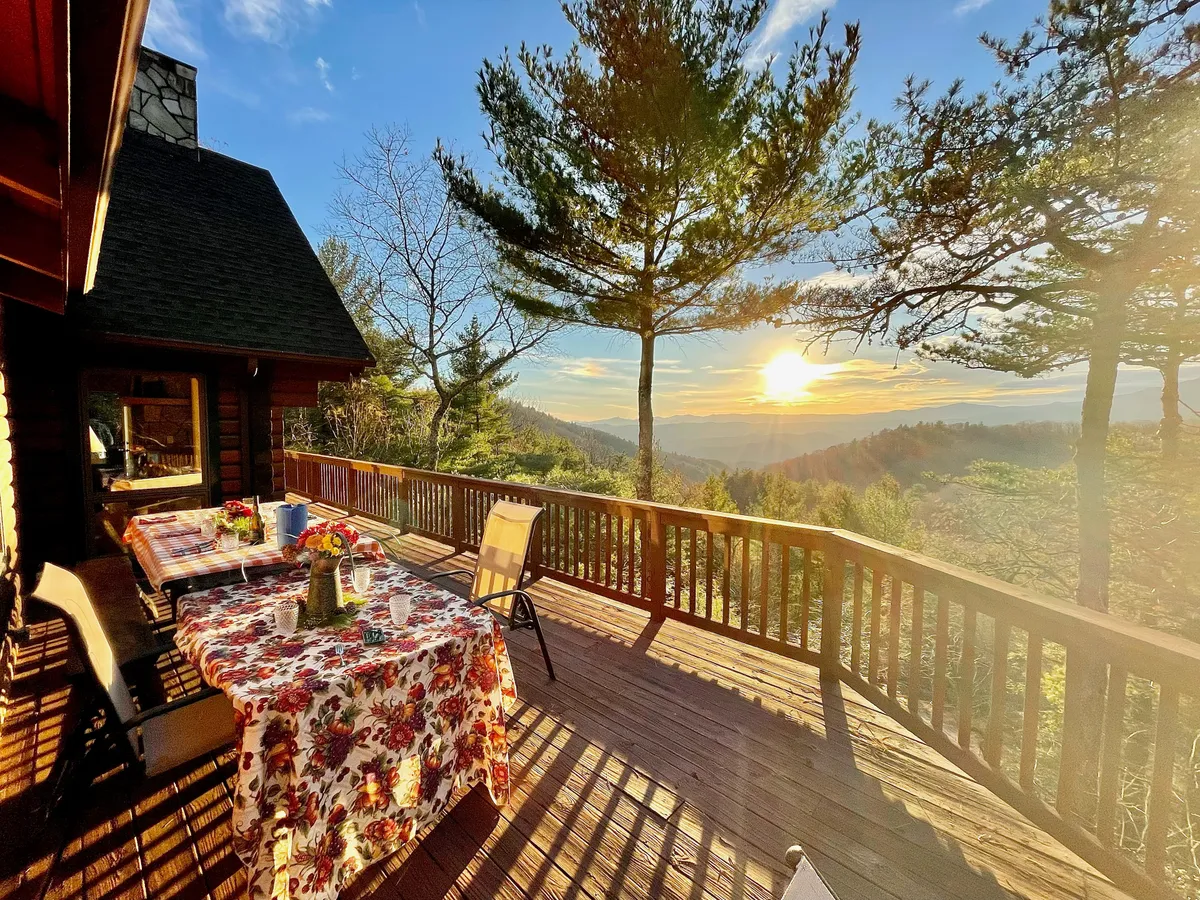 A wooden deck with a dining table dressed in a floral tablecloth is shown at sunset. The scene is surrounded by forest and features a rustic railing, creating a tranquil, inviting setting for al fresco dining and enjoying nature.