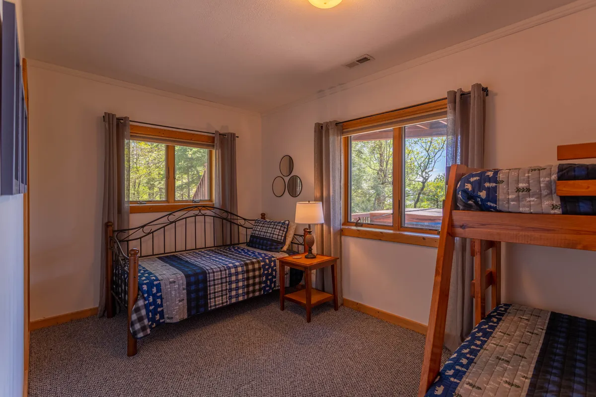 A family-friendly bedroom with a main bed featuring plaid bedding and an adjacent bunk bed. The space includes a side table with a lamp and windows framed by curtains, creating a cozy environment perfect for sleepovers or quiet family moments.