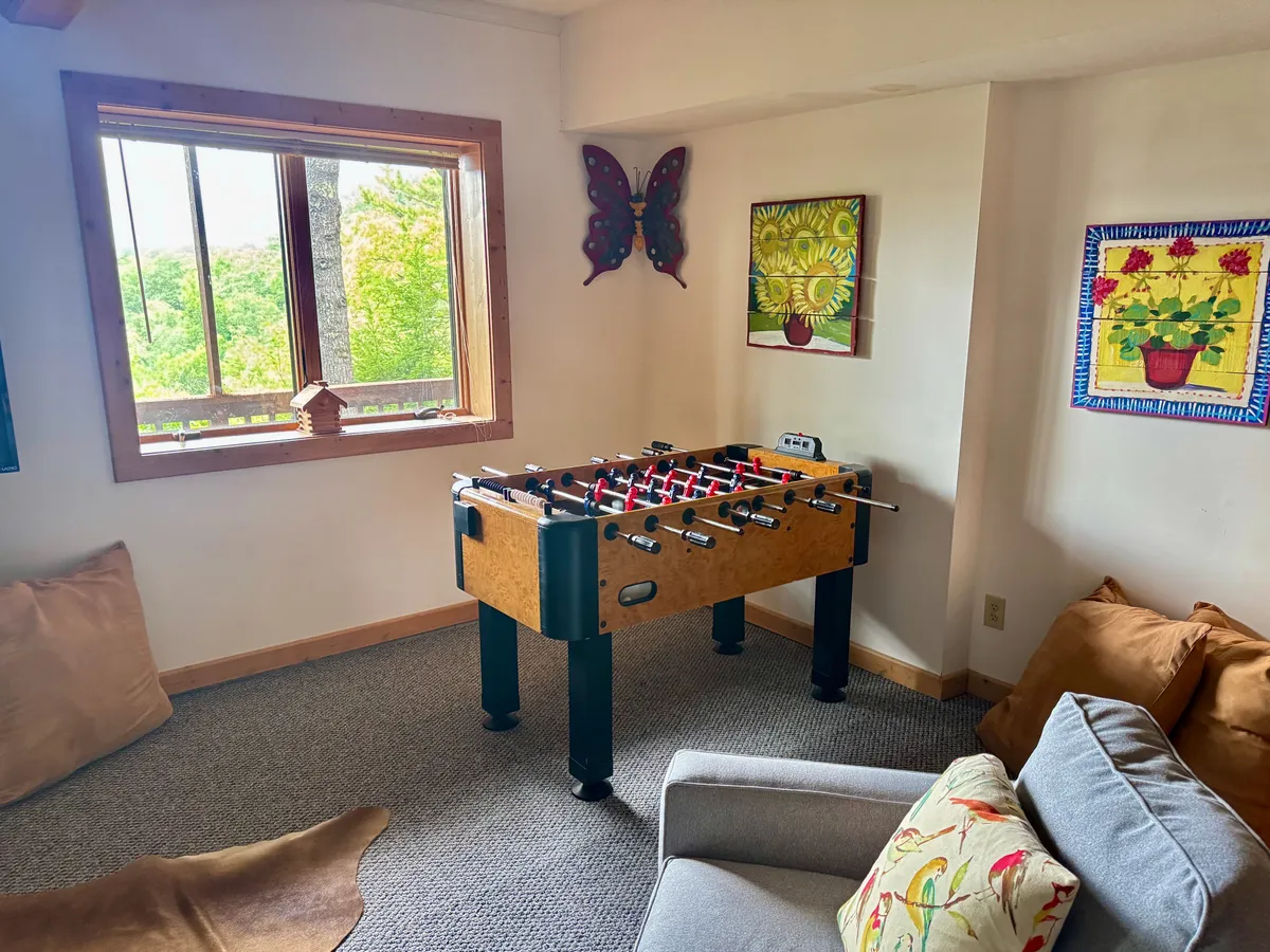 A bright, playful corner with a foosball table front and center, comfortable seating for spectators, and a large window that brings in scenic views. Colorful wall art creates a fun, energetic mood perfect for game nights or friendly matches.