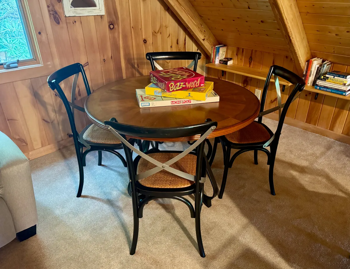 A loft area with a round wooden table surrounded by black chairs, set up for board games and casual conversation. The space is filled with natural light, creating an atmosphere perfect for reading, working, or playful family activities.