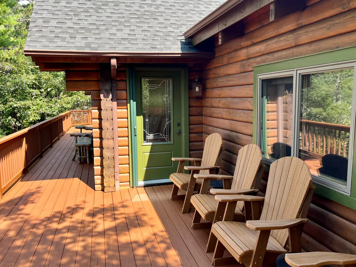 This image captures a portion of the wraparound wooden deck, featuring inviting Adirondack chairs set against a backdrop of lush trees. The green door and large windows of the cabin add a cozy, welcoming feel, while the space is clearly designed for both relaxation and entertaining outdoors.