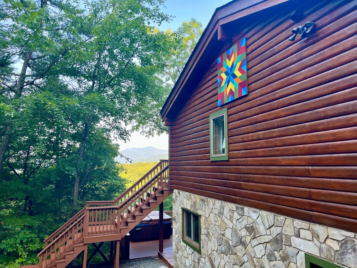 Featuring the stone exterior, a welcoming wooden staircase, and colorful quilt art, this image sets a warm, inviting tone. The home nestles among trees, highlighting both privacy and a strong connection to nature.