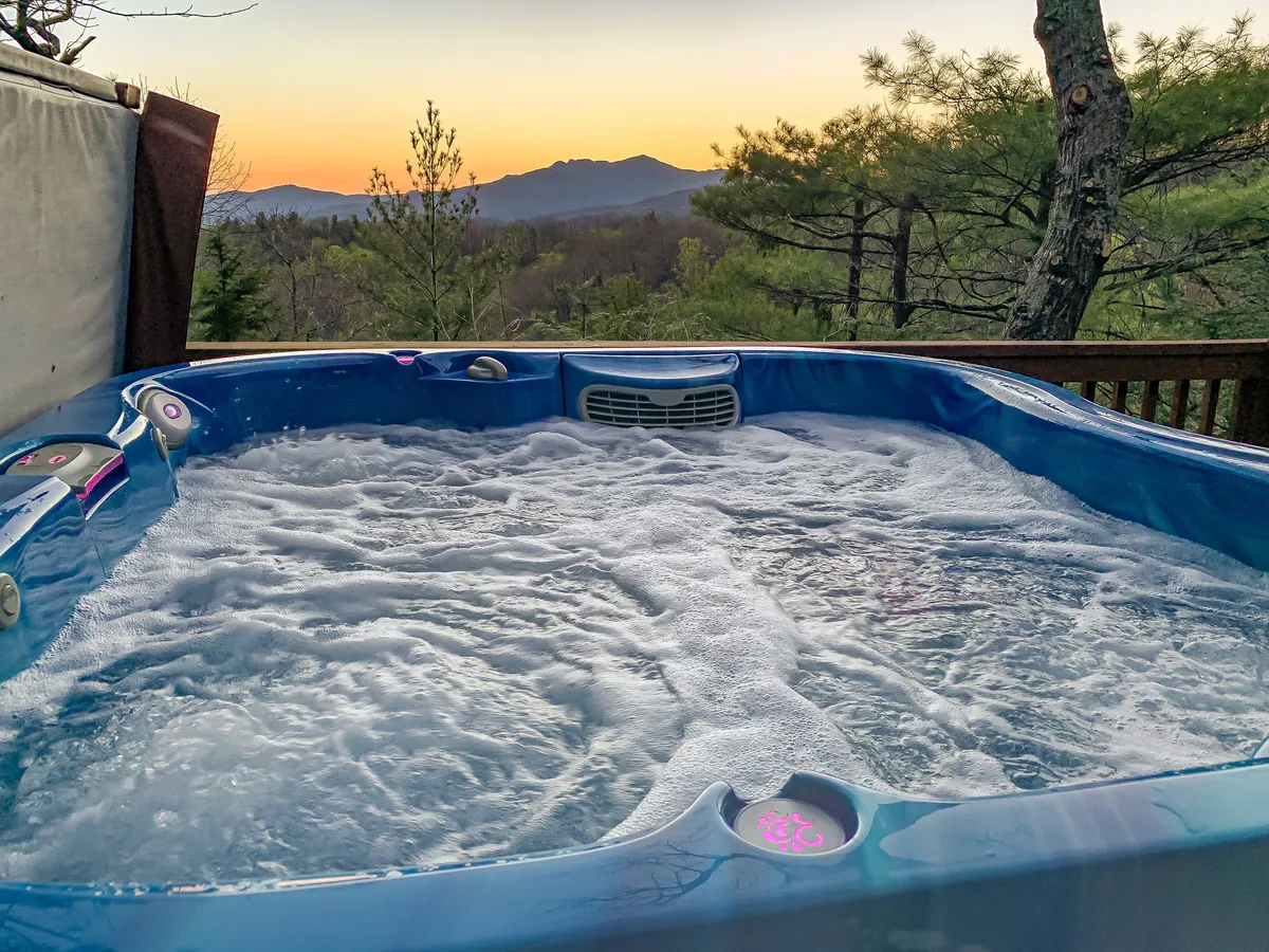 A serene exterior view of a hot tub on a deck overlooking panoramic mountain vistas, with a warm sunset sky painting the scene. The inviting hot tub and ample deck space suggest the perfect place for friends to gather, relax, and soak in the views after an active day.