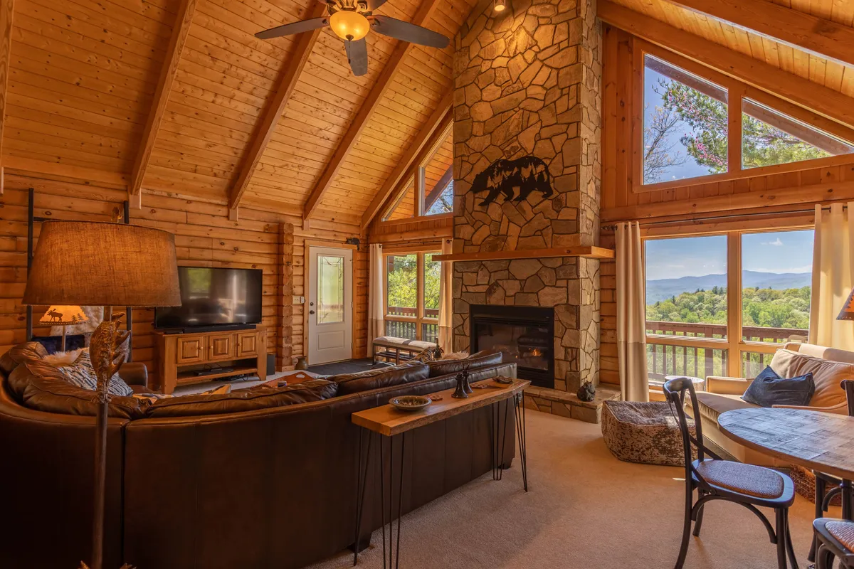 This image showcases the living area with a stone fireplace, large windows flooding the space with natural light, exposed wooden beams, and comfortable seating. The arrangement creates a warm, communal atmosphere ideal for gatherings, relaxation, or enjoying the mountain views outside.