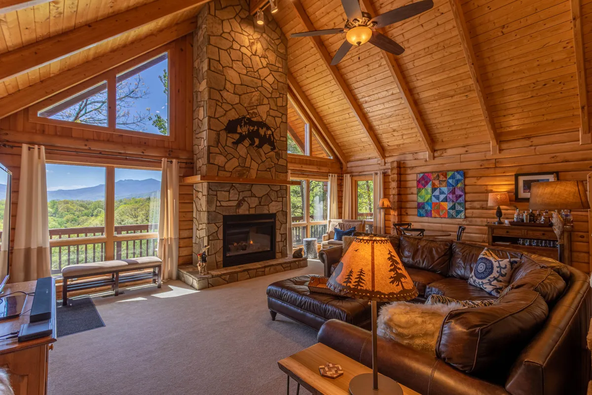 The image showcases a warm and inviting cabin living room with a striking stone fireplace as the centerpiece. Large picture windows frame expansive mountain vistas, while exposed wooden beams and tasteful artwork add rustic charm. A plush leather sofa faces the fire, creating the perfect spot for relaxing, socializing, or soaking in the sunset views with loved ones.