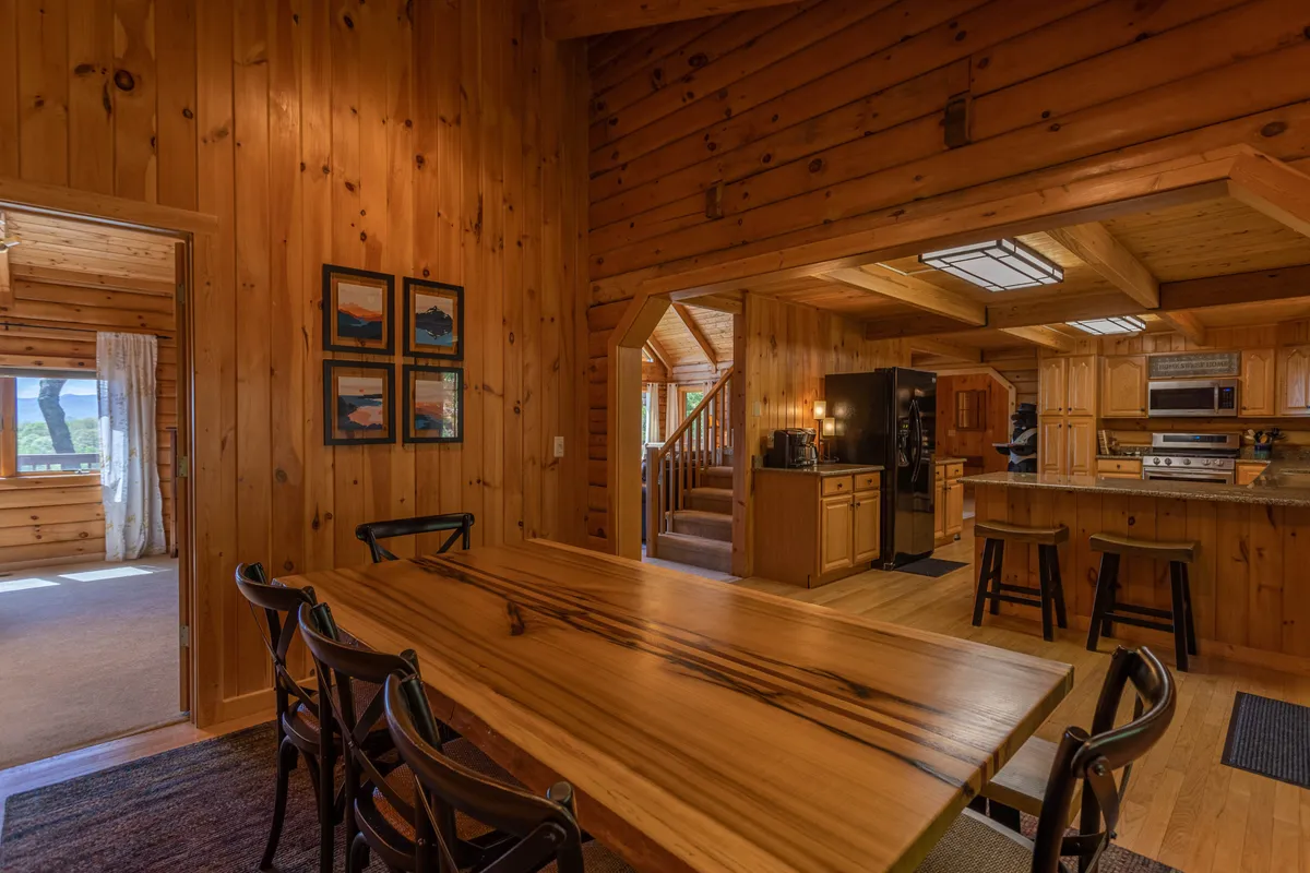 The photo highlights the open-concept layout, featuring a large wooden dining table, exposed wooden beams overhead, a glimpse of the adjacent kitchen area, and art on the walls. The setup conveys a sense of togetherness and is clearly designed for communal dining and socializing.
