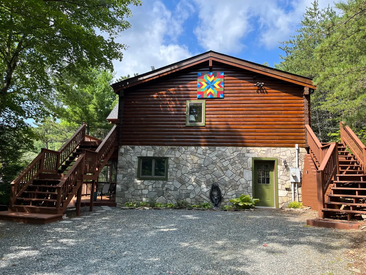 A welcoming exterior shot of Angel’s Envy featuring a stone and wood facade, colorful quilt pattern accent on the wall, staircases leading up to the entrance, and a spacious porch that invites gatherings. The landscape hints at mountain scenery and the feeling of seclusion.